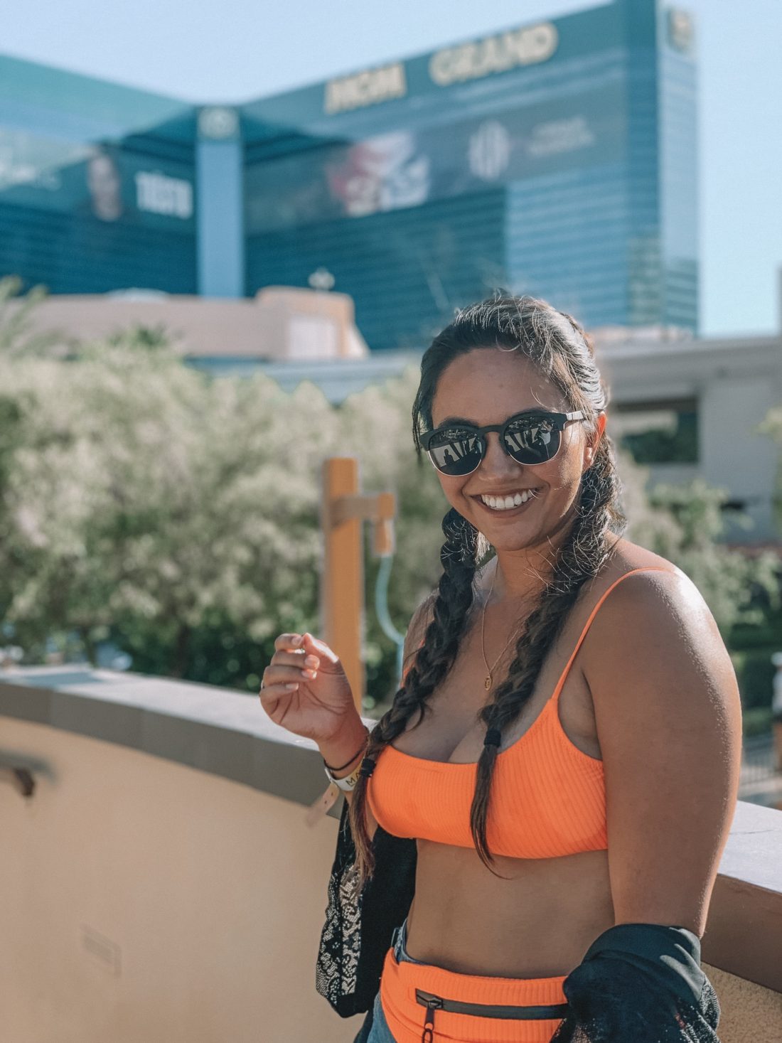 Tan woman in dutch braids standing along a wall wearing a neon orange bikini with a black kimono, denim shorts, and a matching fanny pack