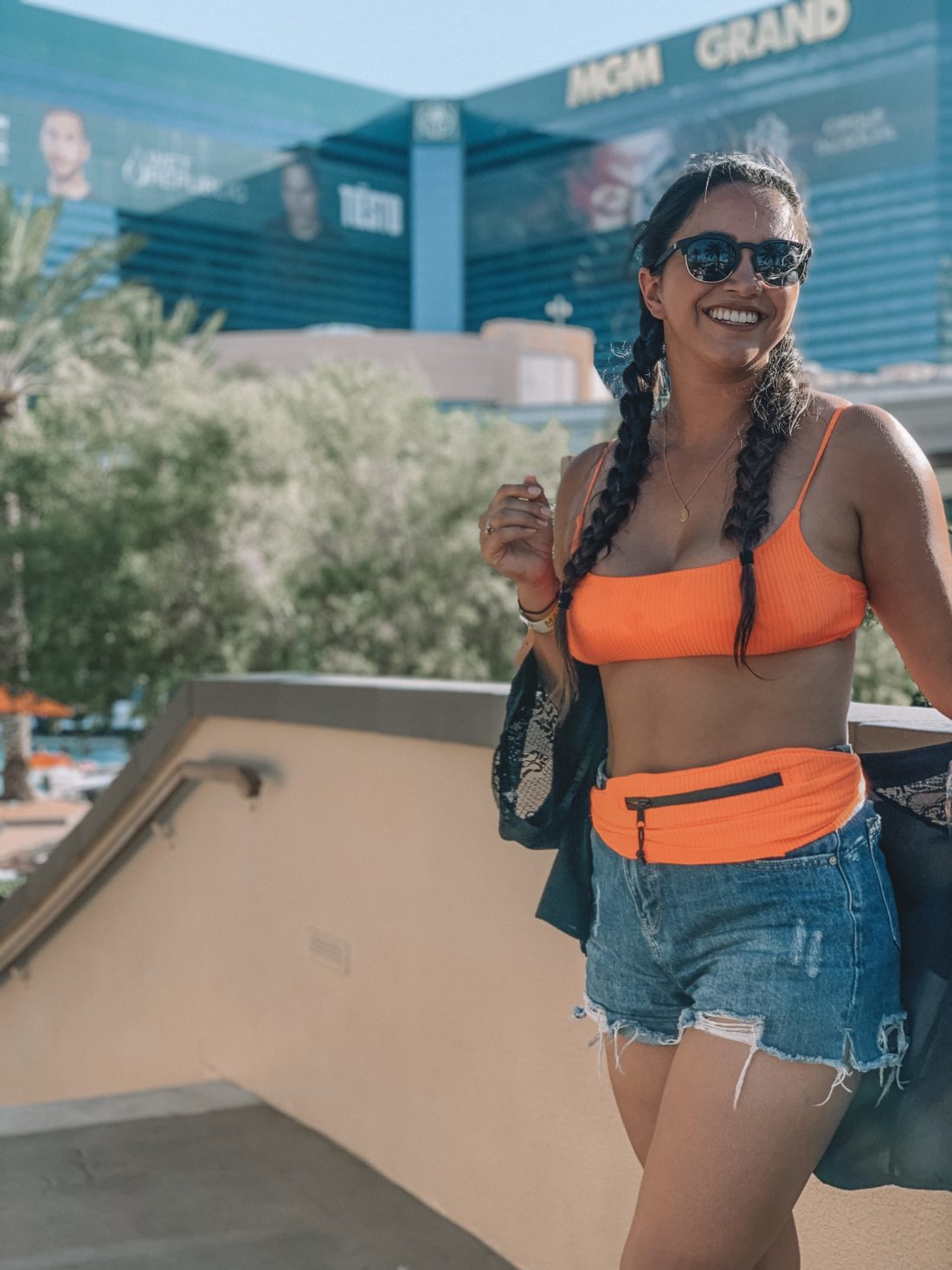 Tan woman in dutch braids standing along a wall wearing a neon orange bikini with a black kimono, denim shorts, and a matching fanny pack