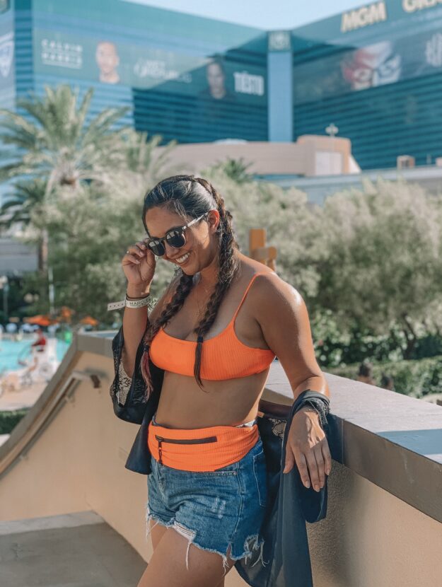 Tan woman in dutch braids standing along a wall wearing a neon orange bikini with a black kimono, denim shorts, and a matching fanny pack